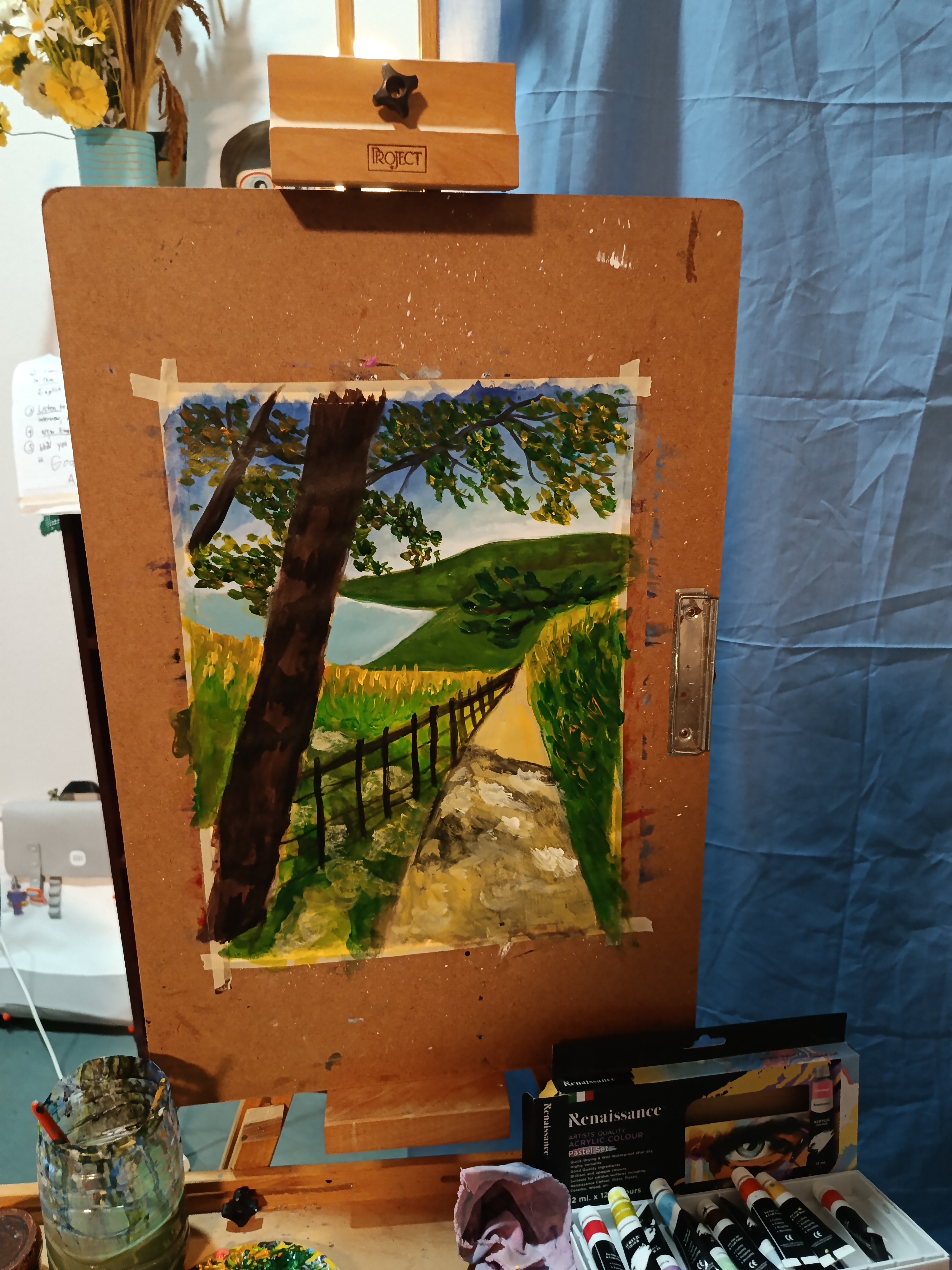 A colorful landscape painting of a narrow path winding through lush green fields. A large tree with spreading branches stands in the foreground, while rolling hills and a calm body of water appear in the distance. The scene is surrounded by greenery beneath a bright, partly cloudy sky